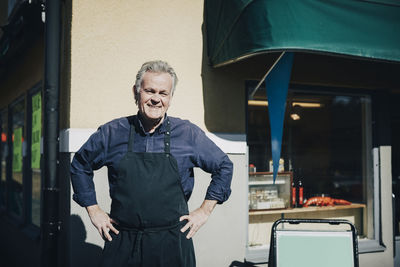 Portrait of smiling male owner standing with hands on hips outside store