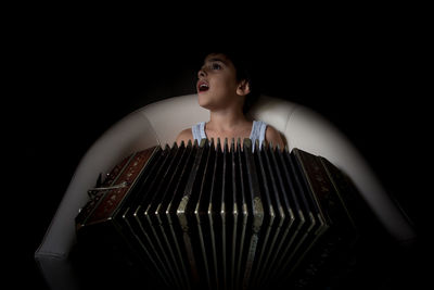 Boy with accordion sitting on chair against black background