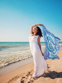 Full length of woman holding scarf on shore against sky at beach during sunny day