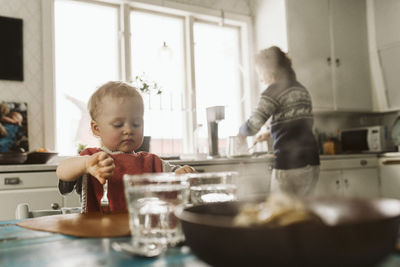 Girl sitting at dining table while mother prepares food in kitchen