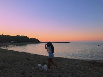 Full length of woman with dog walking at beach during sunset