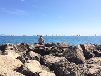 Rear view of people sitting on rock by sea against clear blue sky