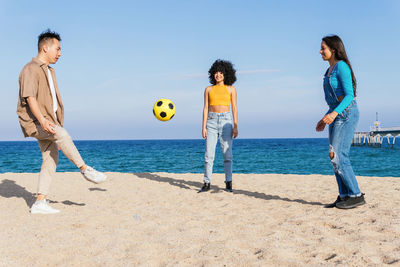 Multiracial male and female friends kicking yellow ball while playing on sandy beach against sea and blue sky on sunny weekend day on resort