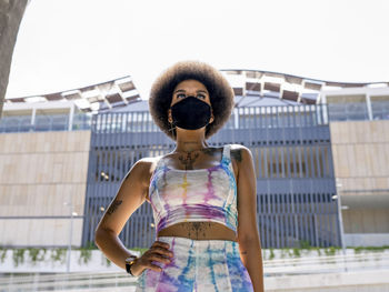 Confident african american female in medical mask and fashionable summer outfit standing in urban area in city during coronavirus outbreak