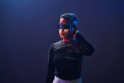 Serious african american female with short hair wearing contemporary vr glasses while standing on purple background in studio with glowing neon lights