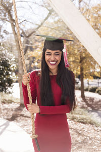 Portrait of laughing young woman wearing mortarboard while holding rope