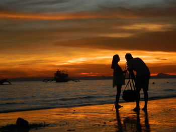 Silhouette couple photographing at beach against sky during sunset