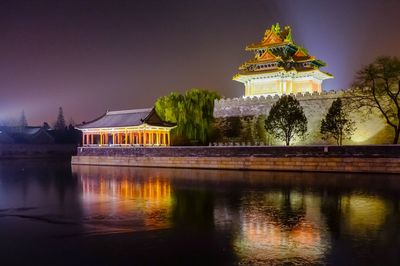 Illuminated building by lake against sky