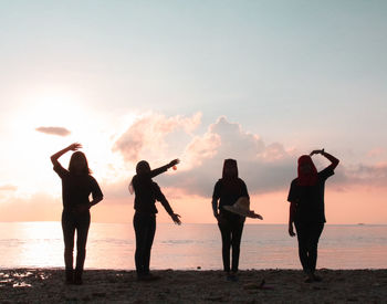 Silhouette friends gesturing while standing at beach against sky during sunset