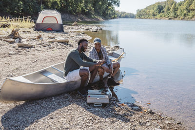 High angle view of friends talking while adjusting fishing tackles on boat at lakeshore