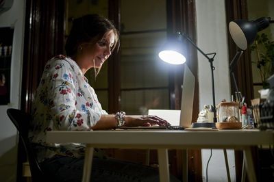 Businesswoman working on laptop by electric lamp in office during night