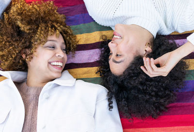 From above of happy multiracial girlfriends with curly hair lying on colorful plaid in park and looking at each other while resting together