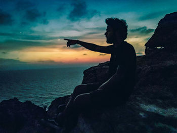 Silhouette man on rock at beach against sky during sunset