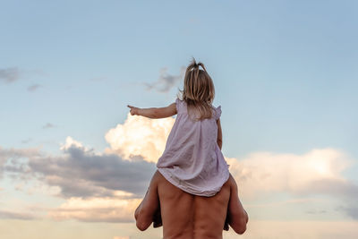 Rear view of man with little daughter standing against sky during sunset