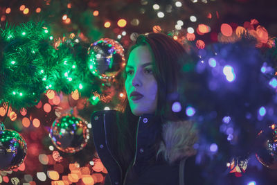 Thoughtful young woman standing against illuminated christmas tree at night