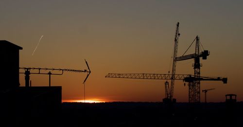 Silhouette cranes at construction site against sky during sunset