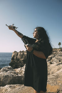 Happy woman with drone looking to the side black dress and remote control oncliff  the sea sunny day