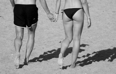 Low section of couple holding hands while walking at beach during sunny day