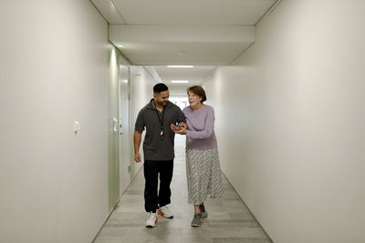 Full length of smiling senior woman walking arm in arm with male nurse at retirement home corridor