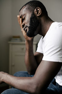 Side view of bearded lonely man covering closed eyes with hand sitting at home