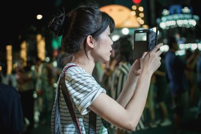 Side view of young woman photographing on mobile phone at night