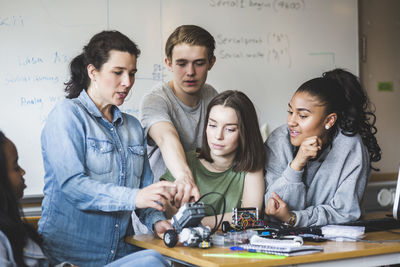 Mature teacher examining robot while sitting amidst students against whiteboard in classroom at high school