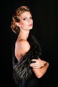 Elegant young woman in black lingerie with fur boa posing at studio on black background