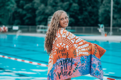 Portrait of smiling young woman wrapped in towel standing by swimming pool