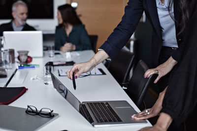 Midsection of businesswomen discussing over laptop with colleagues working in background at office