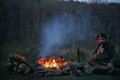 Mid adult man cutting wood by campfire on field at dusk