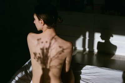 Rear view of shirtless woman sitting on bed