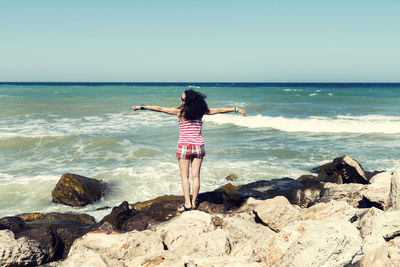 Rear view of woman with arms outstretched standing on rock at beach against clear sky