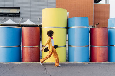 Young woman holding skateboard walking by multi colored pipes