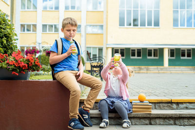 Full length of girl and boy holding drink sitting on steps against school building