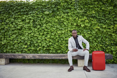 Businessman sitting on a bench with a red suitcase and mobile phone in hand.