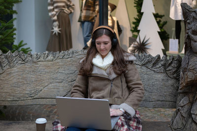 A young woman wearing headphones sitting on a bench and working with a laptop and coffee 