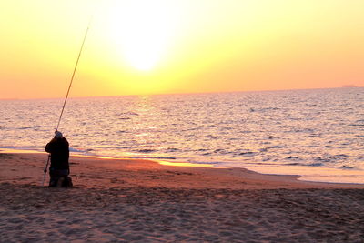 Man fishing on beach against sky during sunset