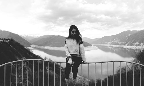 Young woman sitting against mountains and river on railing