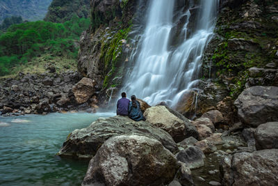 Couple standing at rock in front of waterfall white water stream falling from mountains