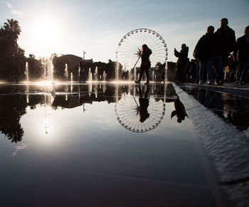 Reflection of people walking by ferris wheel against sky