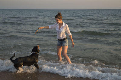 Woman with dog playing in sea during sunset