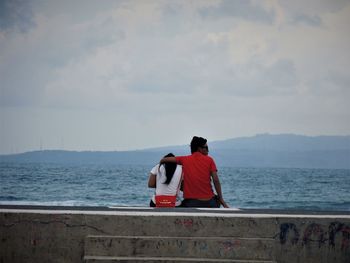 Rear view of men sitting by sea against sky