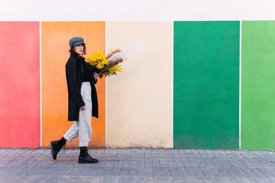 Full body stylish young female in outerwear carrying bunch of fresh flowers and looking at camera while strolling on pavement against colorful wall on city street