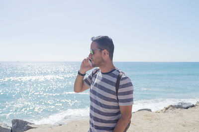 Side view of man listening over phone while standing at beach against clear sky