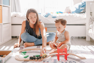 Mother and toddler boy playing with car wooden railway on floor at home. early age education 