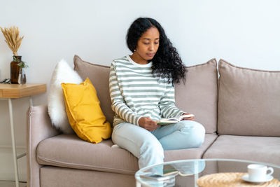 African-american female reading book while sitting on sofa in cozy living room. spending time alone