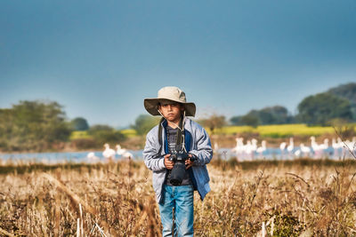 Portrait of boy holding camera standing on field against sky