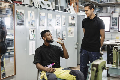 Young man drinking water while looking at coworker in workshop