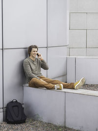 Smiling man sits talks by smartphone on  part of wall. student with rucksack in casual clothes.