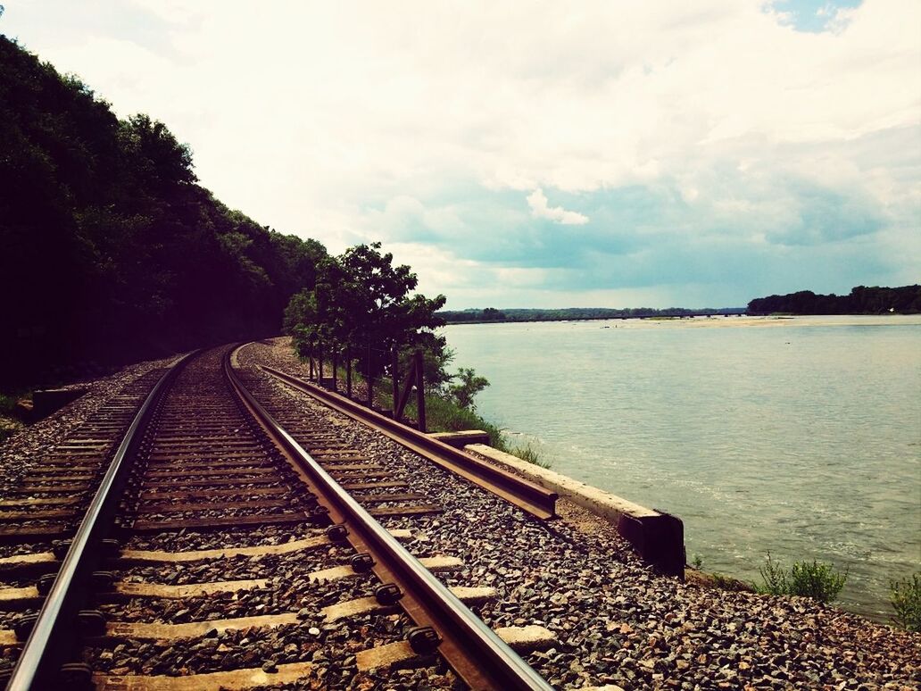 railroad track, rail transportation, sky, transportation, the way forward, water, diminishing perspective, cloud - sky, connection, railing, vanishing point, tranquility, railway track, nature, public transportation, tree, tranquil scene, cloud, high angle view, outdoors